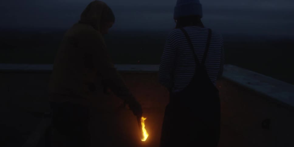 Two people standing in the dark or at twilight are using a burning torch or piece of wood to light something offcamera possibly a fire pit or lantern they are dimly lit by the flames warm orange glow against the deep blue of the sky and the surroun