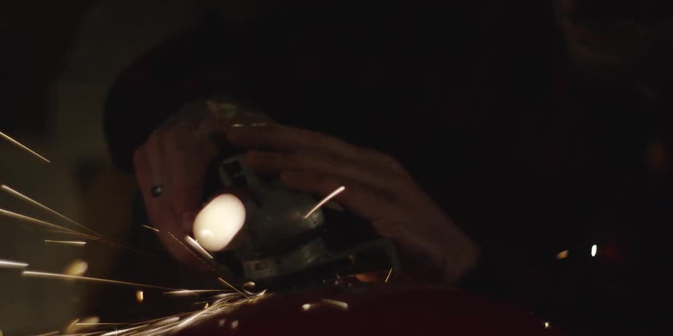 This short video clip shows a person using an angle grinder in a dimly lit environment creating bright sparks as the tool cuts into a red surface the last two seconds feature a closeup shot of the person wearing safety goggles and a beanie
