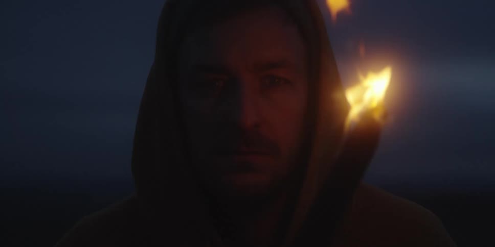 This video shows a medium closeup of a man wearing a hooded garment in a dark setting likely outdoors at night or twilight he is holding a torch which provides the only significant illumination casting a warm flickering light on his face and the sur