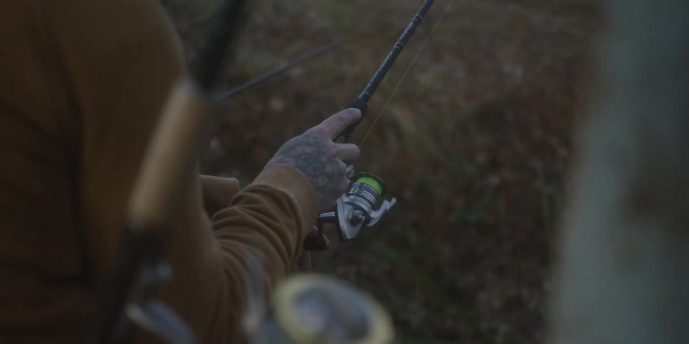 A closeup shot captures the hands of an individual wearing a brown longsleeved shirt and sporting extensive black arm and hand tattoos gripping a spinning fishing rod and reel with the line cast out in a muted natural outdoor setting