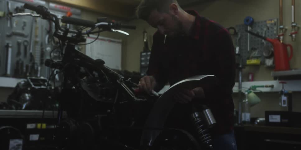 The video shows a man working on a motorcycle in a dimly lit garage or workshop with tools visible on the walls behind him he is focused on the central part of the motorbike perhaps performing repairs or modifications