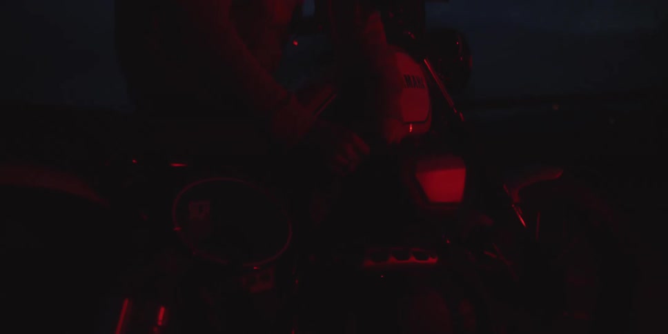 In the deep twilight a hooded man leans intently over his motorcycle making adjustments as the scene is cast in an intense red glow