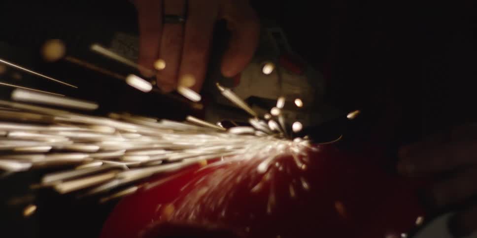 A person uses an electric angle grinder on a rounded red object creating a bright intense shower of whitehot sparks in a dark highcontrast environment