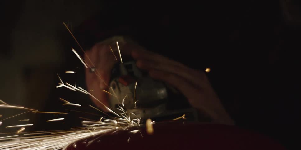 A person in the dark operates an angle grinder on a curved red metal surface sending bright long sparks flying outwards the sparks momentarily illuminate the hands gripping the power tool and the surrounding gloom