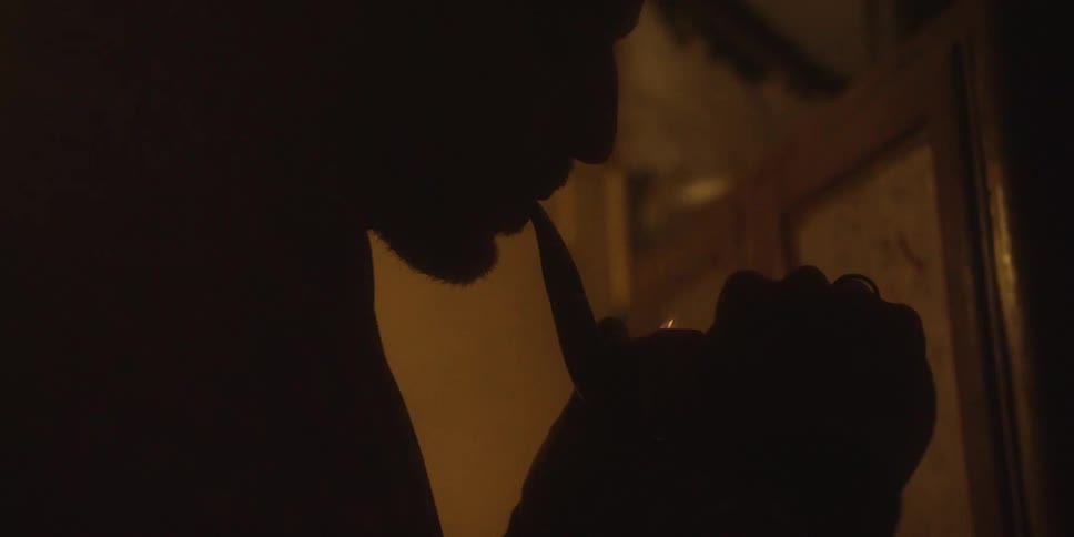 In a dark warmly lit room a mans silhouette is briefly illuminated by the flickering flame of a lighter as he lights his smoking pipe