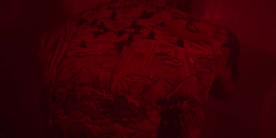 A closeup shot under intense red lighting shows the back of a person focusing on a large intricate tattoo covering their entire back the tattoo features elements like a flock of birds mountains text that says of narrow roads and a prominent nucle