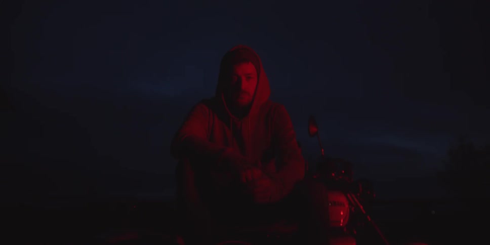 A man wearing a hooded sweatshirt is sitting on or next to a motorcycle in the dark illuminated primarily by a deep red light sourcethe lighting creates a moody silhouettelike effect with the man looking directly towards the camera as the video slo