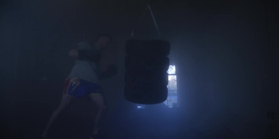 A young man punches a stack of tires during exercise inside a dark room as faint sunlight streams through a nearby window