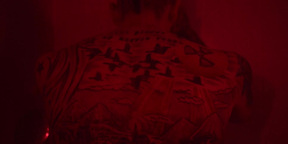 A man with a heavily tattooed back is shown in closeup dimly lit by a pervasive intense red light making the scene feel like a darkroom or a heat lamp the tattoos include flying birds mountains and a large nuclear symbol