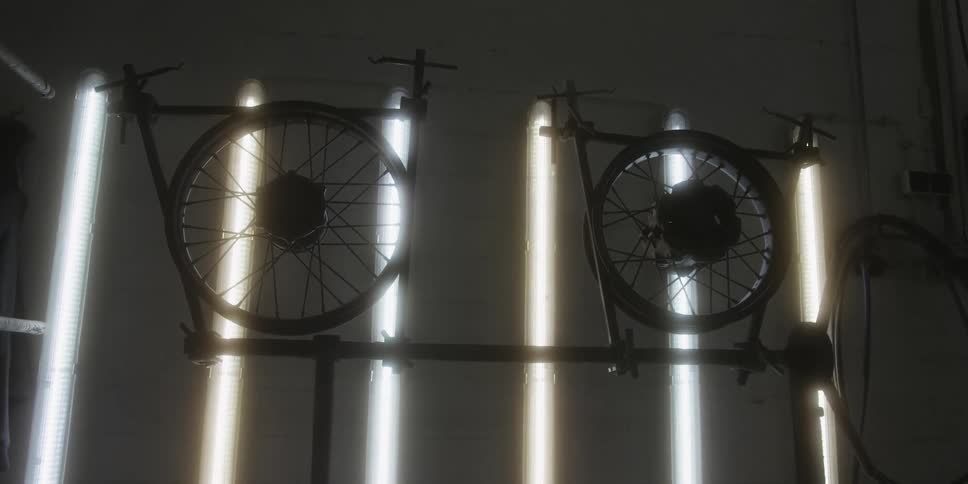 Two bicycle wheels and their frames are silhouetted against a dark background brightly illuminated by vertical fluorescent light tubes in an indoor setting