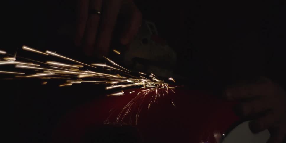 In a dark workshop a worker uses an angle grinder on a curved piece of red metal sending a brilliant shower of sparks into the blackness