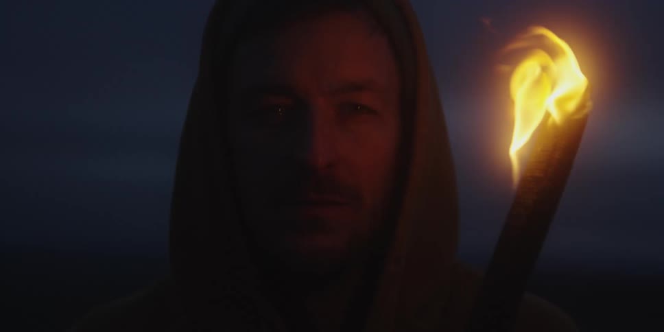 A hooded man holds a blazing torch that dramatically lights his face in the dark