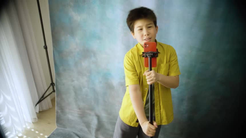 Young boy wearing a white shirt singing and playing air guitar with a tripod holding a smartphone on a photography studio indoors during day time
