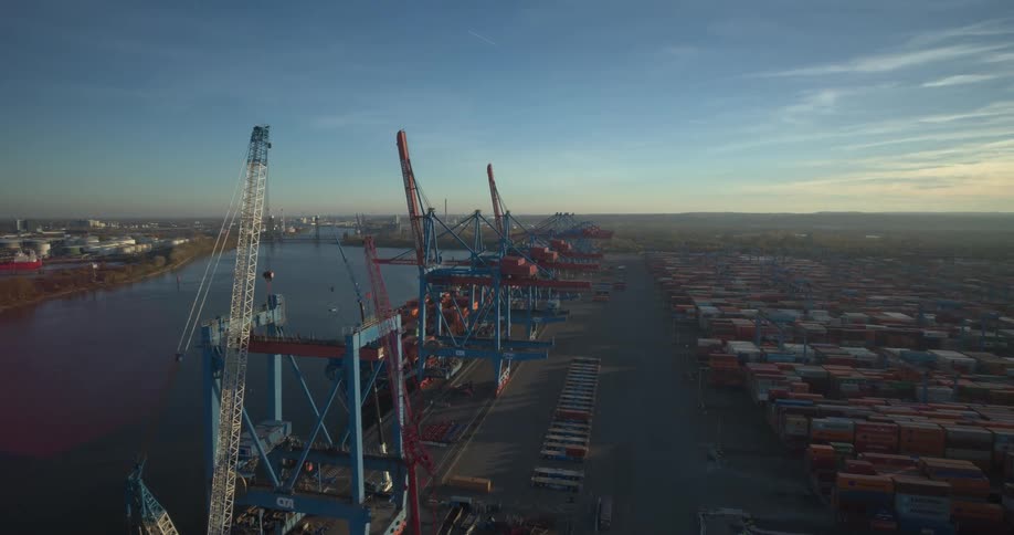 An expansive aerial view captures a large active container port situated along a river showing numerous blue and red gantry cranes beside endless rows of stacked cargo containers under a bright sky