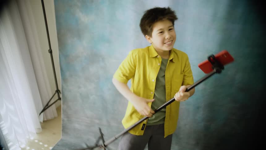 Teenager boy playing air guitar and singing into a tripod with a smart phone hanging from it on a photography studio outdoors during day time