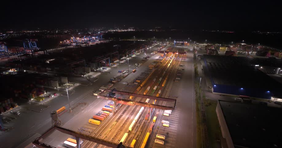 A wide high angle aerial view shows a brightly lit cargo depot with rows of shipping containers and trains at night