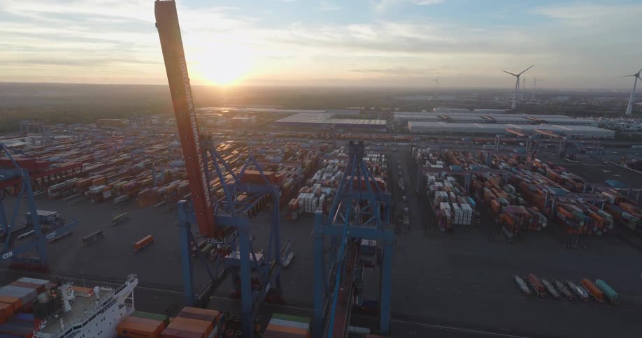 A sprawling shipping container terminal with towering cranes and stacked cargo is bathed in the golden light of sunset