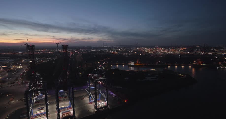 Container cranes lit up operating in industrial port at dusk with wind turbines in the background