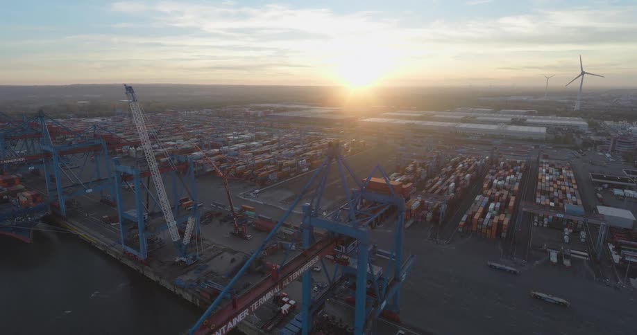 A high angle aerial view shows a large industrial container terminal with multiple red and blue port cranes a docked ship rows of shipping containers and distant wind turbines under a bright sunrise
