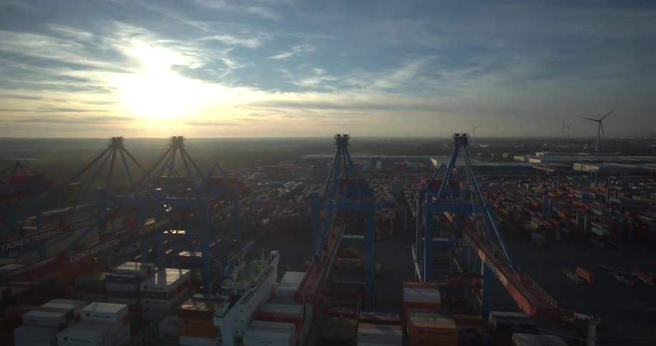 A high angle aerial view shows a massive container terminal at sunset with large blue port cranes a moving white crane a cargo ship docked and rows of shipping containers