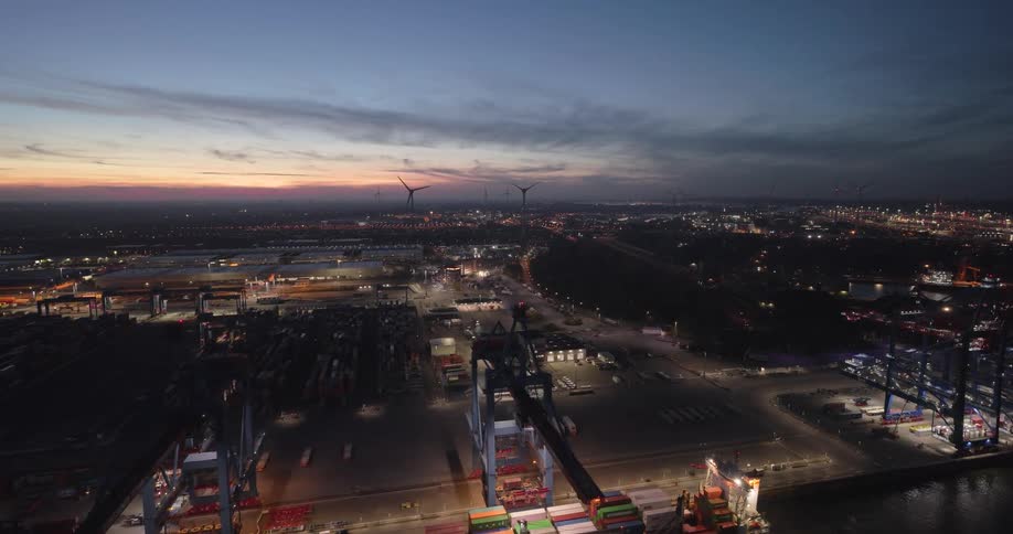 Industrial port lit at dusk with cargo ship docked and wind turbines in the background