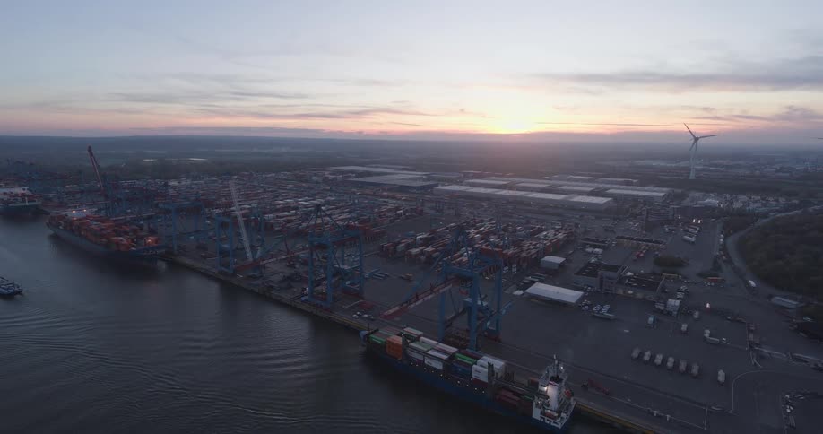 Ships at a loading dock with cranes on the coast and containers stacked on the ships and on shore as wind turbines spin in the distance
