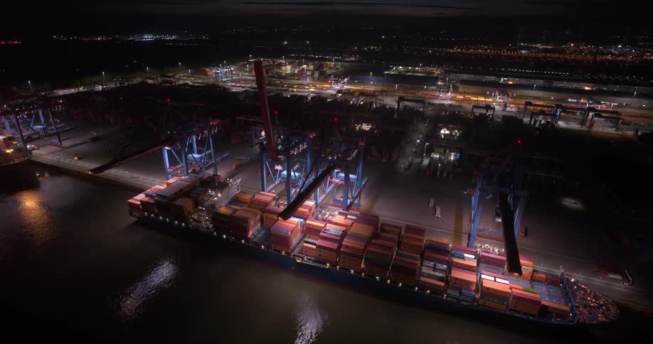 Lights on a loading dock at night as cranes hang over boats filled with shipping containers