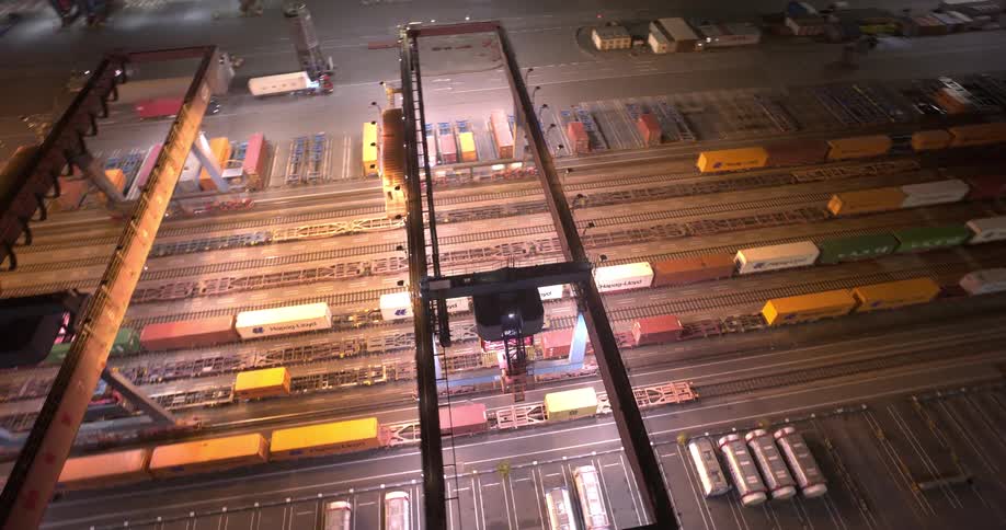 An aerial view circles a massive brightly lit shipping port at night revealing gantry cranes moving over long trains loaded with colorful containers