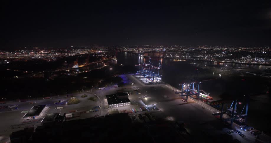 Aerial view of a brightly lit industrial port with cranes and stacked containers along the water at night