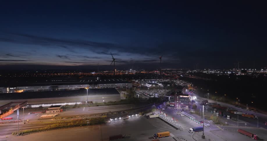 Nighttime aerial view shows an illuminated industrial park with wind turbines in the background