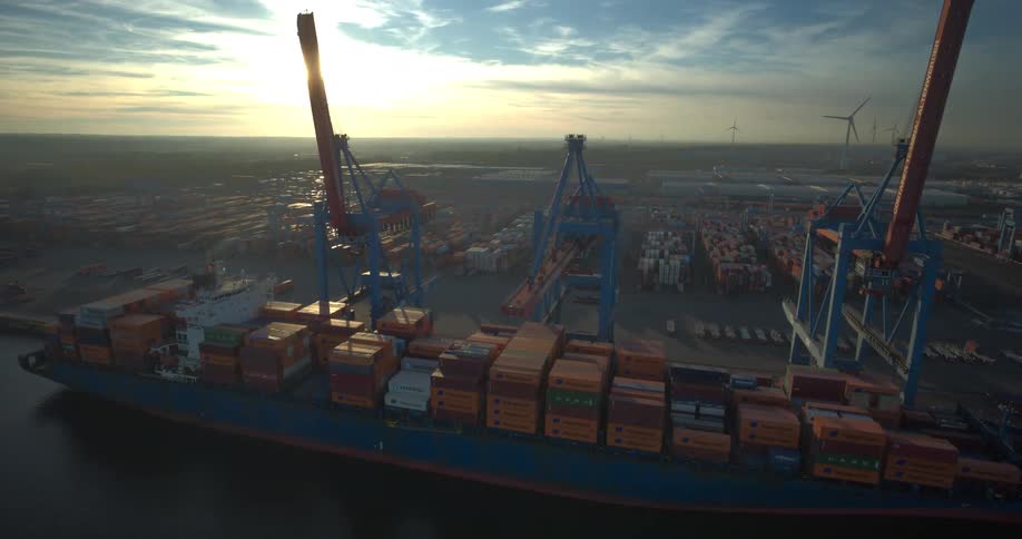 Cargo ship full of containers docked on a harbor next to cranes outdoors during dusk or dawn