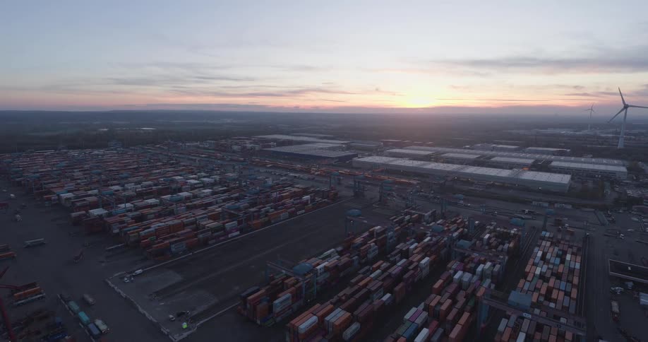 Ships at a loading dock with cranes on the coast and containers stacked on the ships and on shore as wind turbines spin in the distance