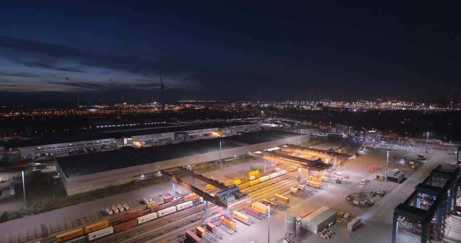 Semi trucks drive through a shipping center located on the edge of a brightly lit city at night