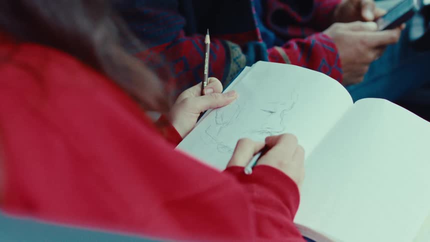Female person wearing a red sweater sketching a portrait of a man sitting in front of her using a smartphone indoors during day time