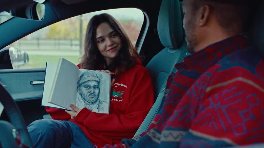 A woman wearing a red sweatshirt sits in the driver seat of a car showing a drawing in a sketchpad to the man in the passenger seat