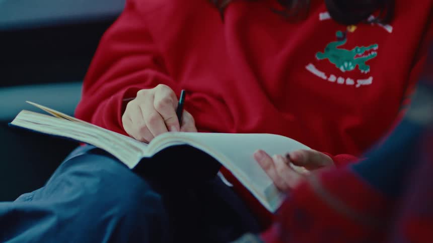 A close view of a woman wearing a red sweatshirt writing with a black pen in an open white notebook resting on her lap in a blurred dark setting