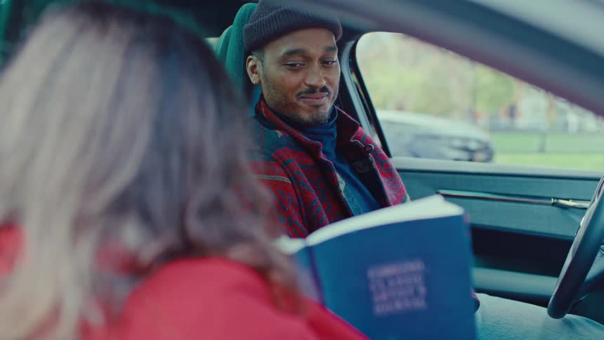 Young adult man wearing a beanie sitting inside a car and opening his eyes to be surprised and laugh at a book that a woman is showing him outdoors during day time