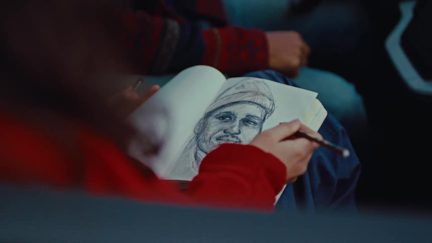 A woman sketches a portrait of a person in a knit cap in a spiral bound notebook