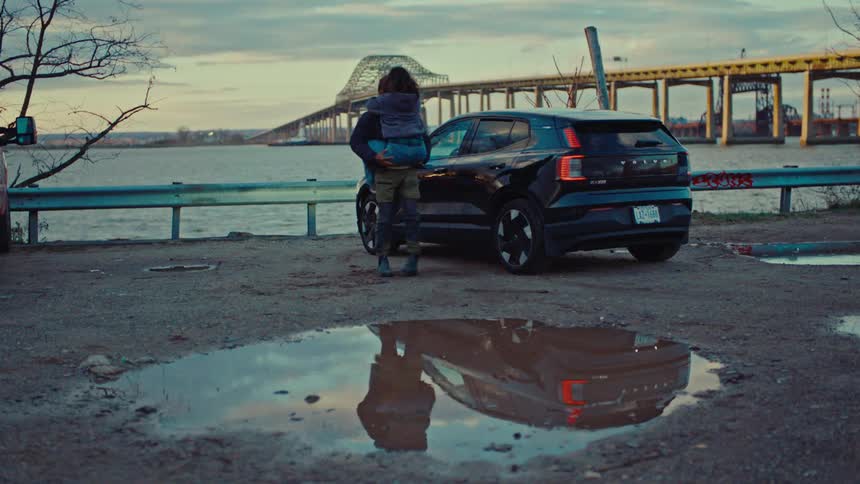Couple embracing beside black electric vehicle parked by waterfront with bridge in the background