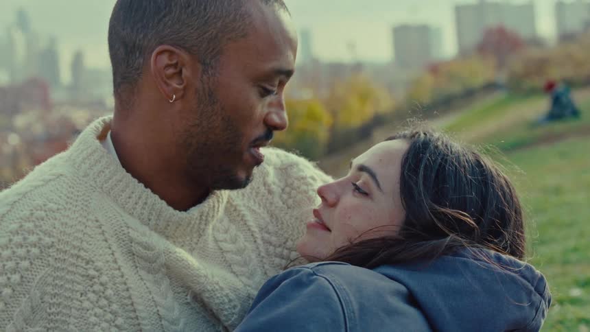A man and a woman in winter clothing embrace and smile in a grassy park setting with an urban skyline blurred in the background