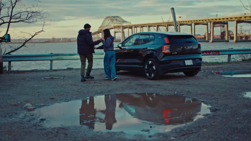 Couple dancing on wet ground beside black suv parked near water with bridge in the background