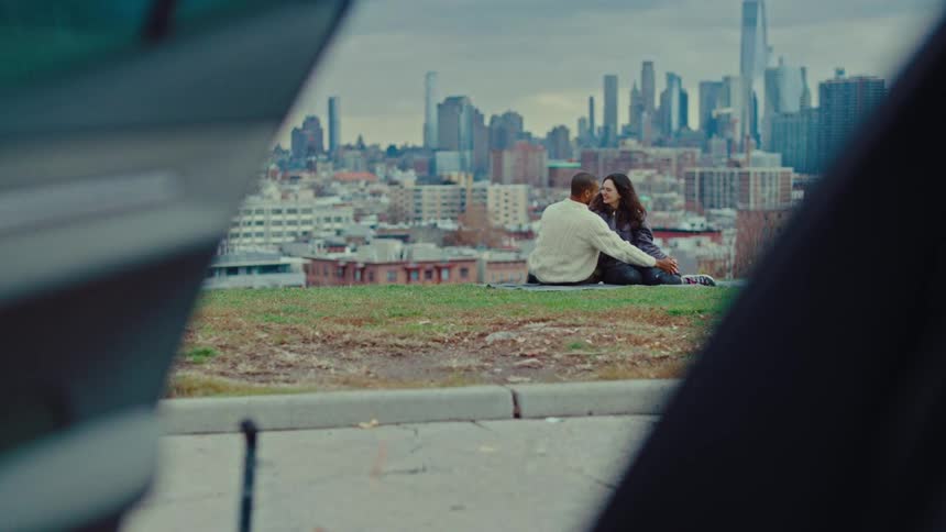 Couple sits on the ground with the city skyline in the background while a car sits with its door open on the nearby road