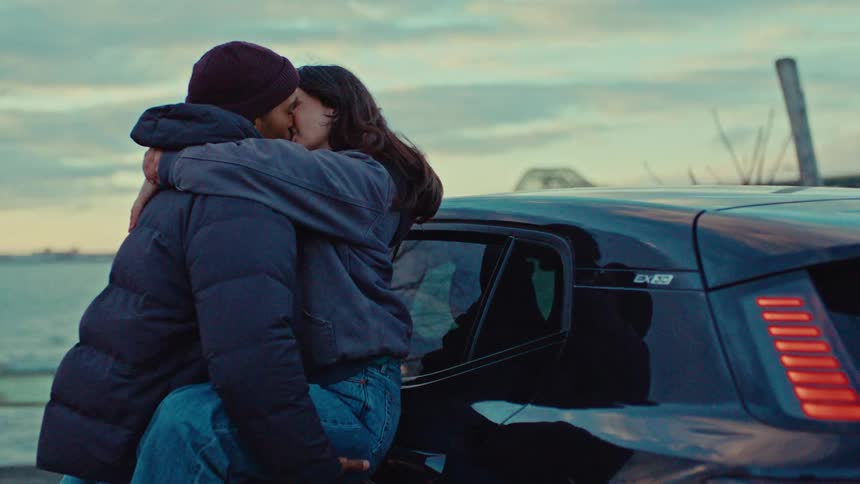 A young couple shares an intimate moment beside a parked car the man lifting the woman into his arms as they embrace and kiss passionately against a cool overcast waterfront backdrop
