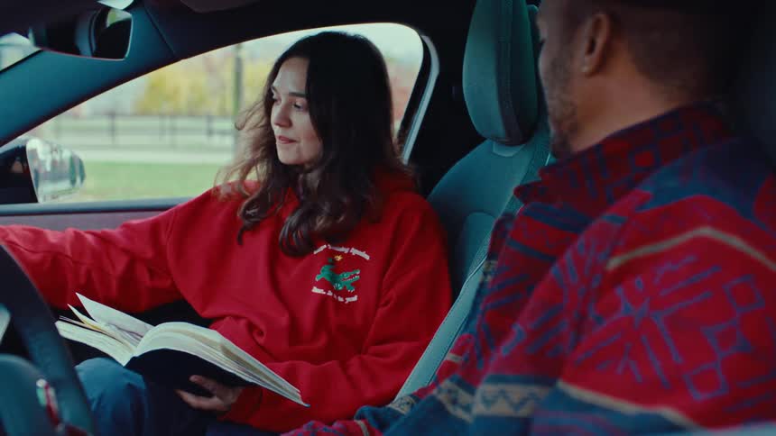 Sitting behind the wheel of a parked car a woman in a red sweatshirt turns her sketchbook around to proudly show a pencil portrait to her male companion smiling as she shares her art