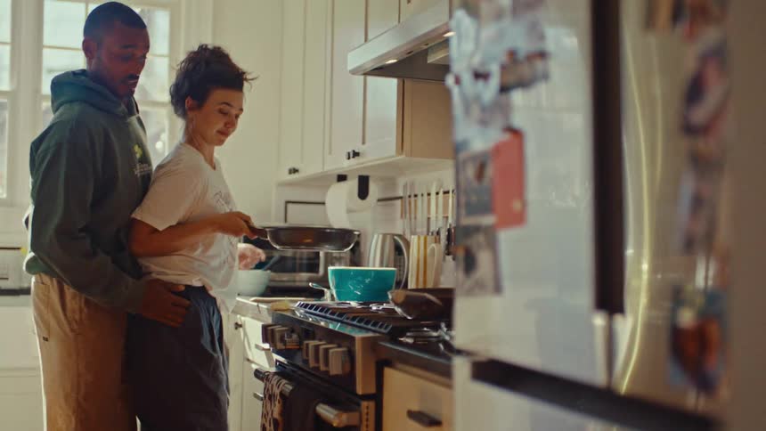 An affectionate couple cooks together in a sunlit kitchen beside the stove