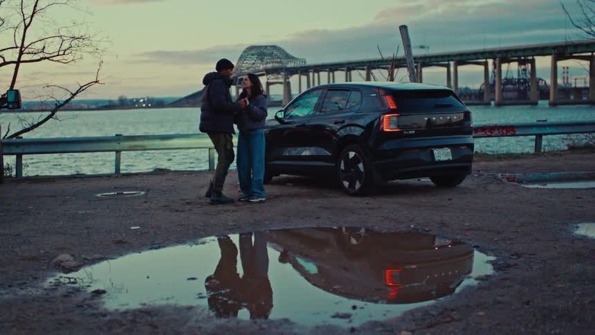 A couple dances holding hands next to a black suv by a river at dusk with a bridge in the background and the car reflected in a puddle