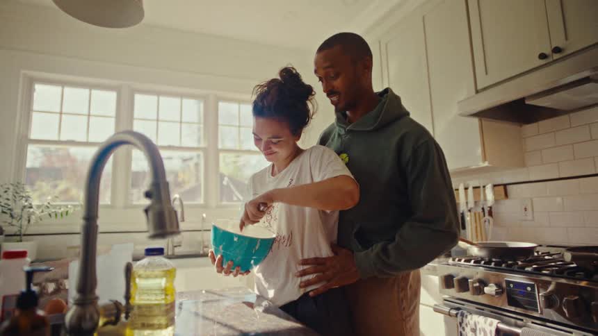A happy couple embraces in a sunlit kitchen while the woman whisks ingredients in a blue bowl