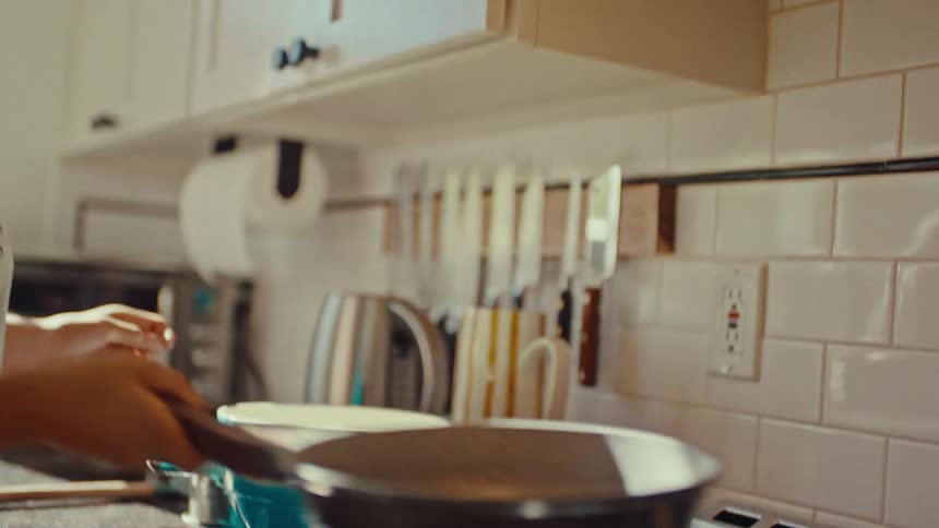 Young adult woman flipping a pancake on a pan on top of a stove with a young adult man hugging her from the back while both smile in a kitchen indoors during day time