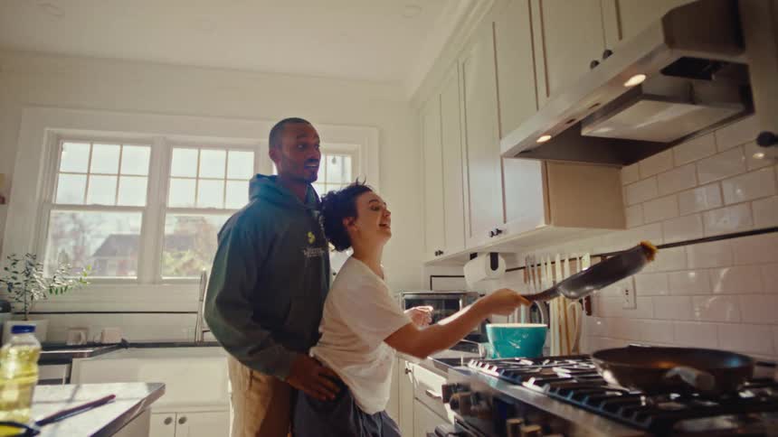 A man embraces a laughing woman from behind as she playfully flips food in a frying pan