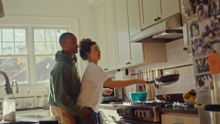 Couple in a kitchen while the person in front tosses food upward while flipping the frying pan on the stove